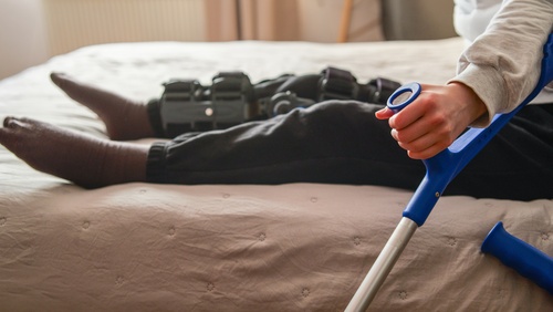 A person wearing a knee brace and holding a crutch sits on a bed, indicating recovery and rehabilitation after an injury or surgery