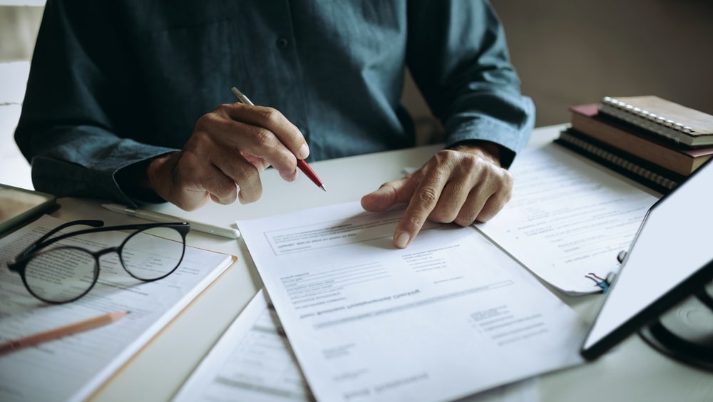 Person reviewing paperwork with a pen at a desk covered with documents